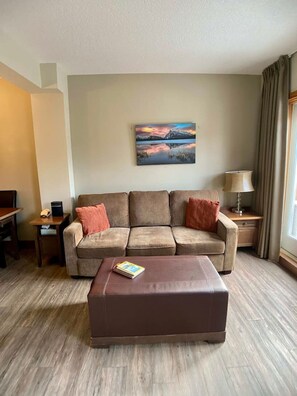 Un salon avec un canapé, une table basse, un tableau au mur, une lampe et une fenêtre avec des rideaux.