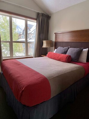 Une chambre d’hôtel avec un grand lit, une tête de lit en bois, une table de chevet avec une lampe et une fenêtre avec des rideaux.