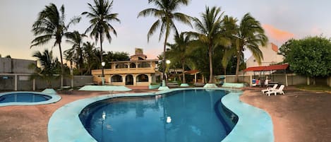 A swimming pool with a curved design, surrounded by palm trees and a building with a red roof.