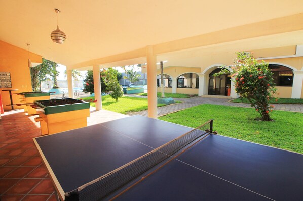 Área externa coberta com mesa de pingue-pongue, mesa de futebol de mesa e mesa de bilhar. Há plantas verdes e um pequeno jardim.