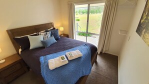 A neatly made bed with a blue quilt and pillows, a wooden headboard, and a nightstand with a lamp. - Feathertop Views and Barn (Smoko)