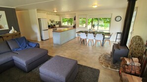 A modern living room with a grey sectional sofa, a small ottoman, and a dining area with a wooden table and white chairs. A kitchen with white cabinets and a refrigerator is visible through an open door. - Feathertop Views and Barn (Smoko)