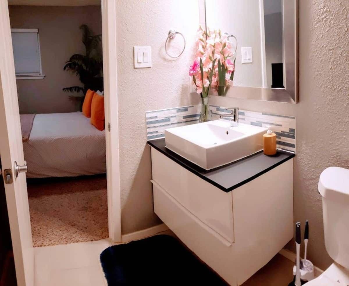 Ein Badezimmer mit weißem Waschbecken, Spiegel und Toilette. Auf dem Boden liegt ein kleiner Teppich und in der Ecke steht eine Pflanze.