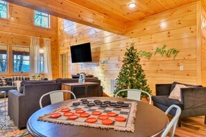 L’intérieur chaleureux d’un chalet, avec un décor en bois, un sapin de Noël, un téléviseur à écran plat et un jeu de société à carreaux posé sur la table.