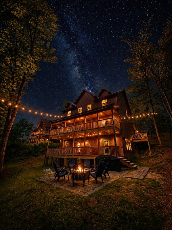 A two-story wooden cabin with a balcony, string lights, and a fire pit.