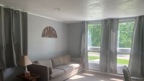 A living room with a sofa, a lamp, a chair, and a window with curtains. - Killington / Rutland 1 Bdroom Cabin, Cabin #1 (Mendon)