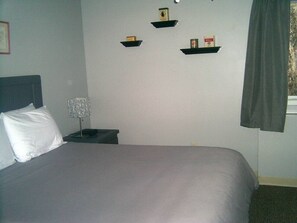 Un lit avec une couvre-lit grise, une table de chevet avec une lampe et trois étagères au mur.