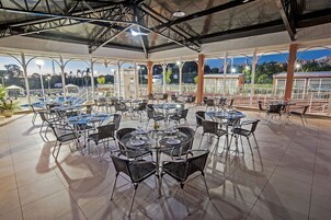 A spacious outdoor dining area with multiple tables and chairs, large windows offering views of the cityscape, and a covered roof structure.