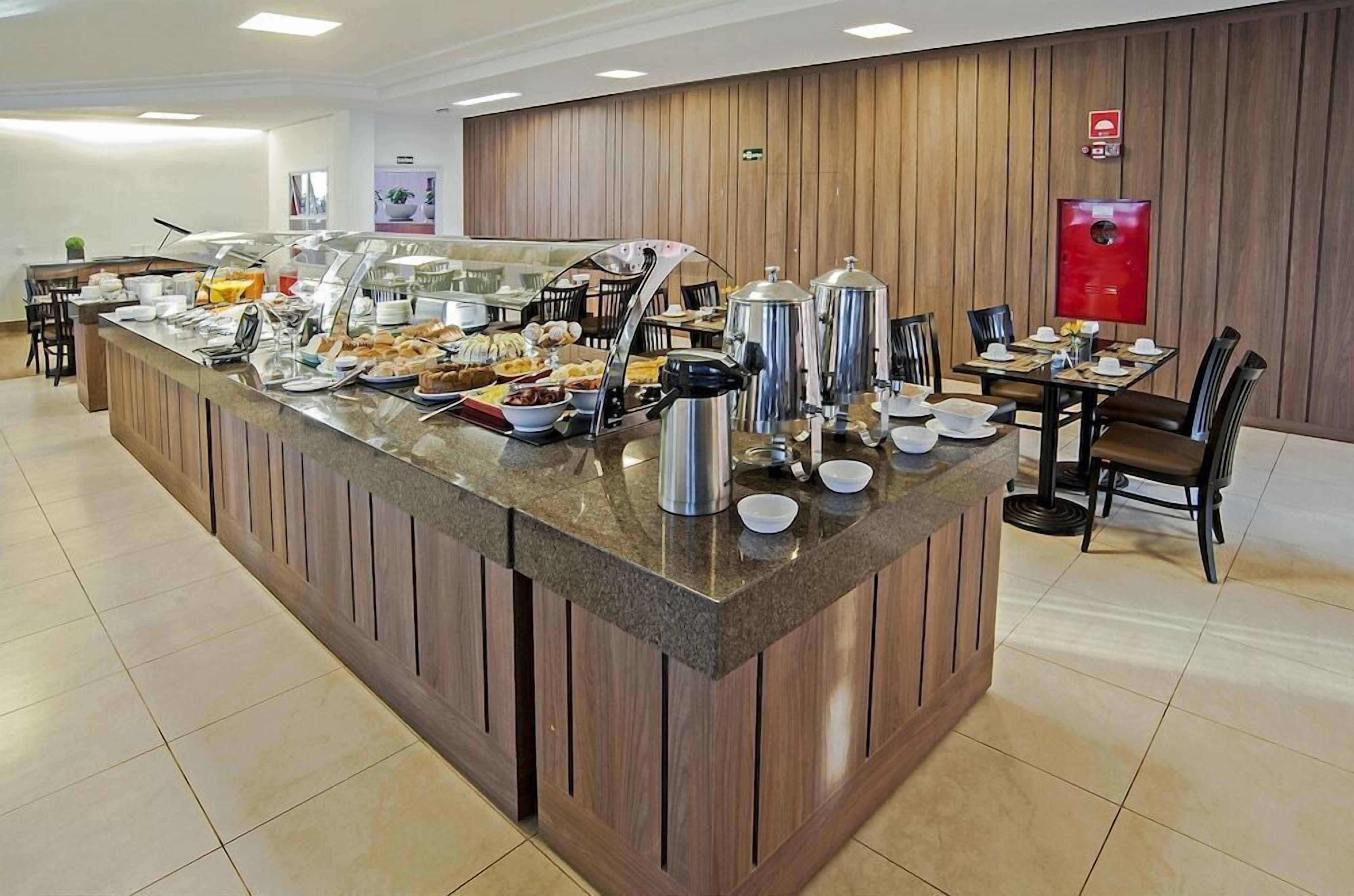 A buffet area with a variety of food items, including fruits, pastries, and beverages, with a wooden panel wall and a fire extinguisher mounted on it.