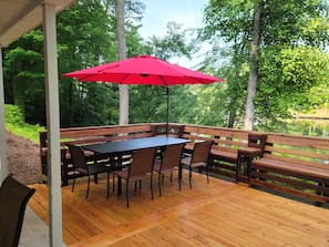 A wooden deck with a table and chairs, a red umbrella, and a view of a lake surrounded by trees.