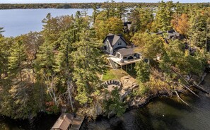 An aerial view of a lakeside property with multiple houses, a dock, and a stone fireplace. - Stunning 5 + Bed, 3 Bath Cottage on the South End of Lake Muskoka! All Day Sun! (Gravenhurst)