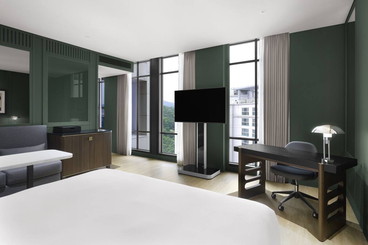 A modern hotel room with a large bed, a desk, a chair, a television, and a view of the city through large windows.
