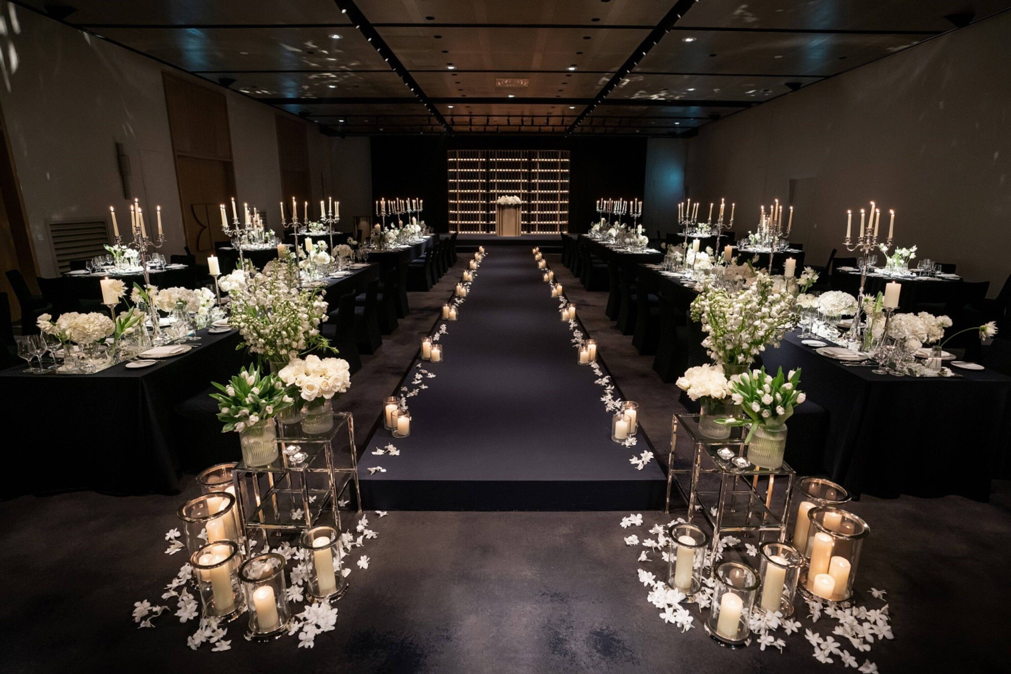 A long, narrow room with tables set for a formal event, featuring candle-lit centerpieces and a dark carpeted floor.