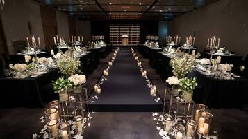 A long, narrow room with tables set for a formal event, featuring candle-lit centerpieces and a dark carpeted floor.