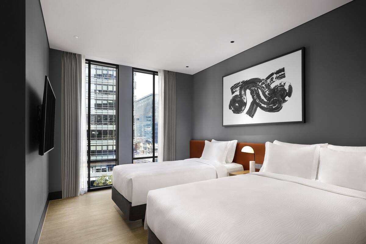 A hotel room with two beds, a large window with a city view, a mounted TV, and a framed abstract artwork on the wall.