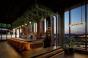 A modern bar area with a long counter, seating, and large windows offering a city view.