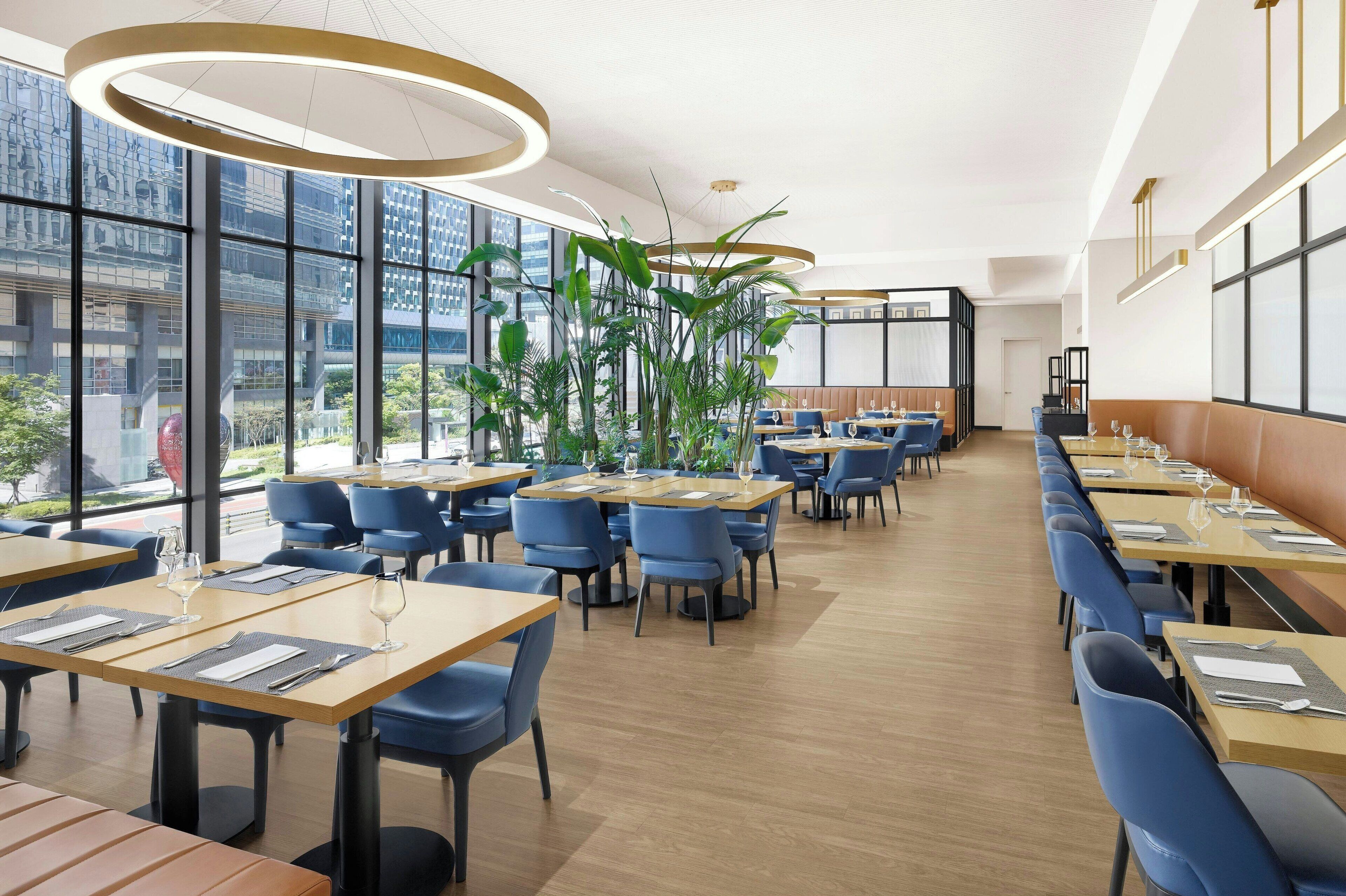 En moderne restaurant med lange borde, blå stole og store vinduer med udsigt over byen.
