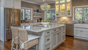 A modern kitchen with white cabinetry, a stainless steel refrigerator, a large island with a bowl of fruit, and a hanging light fixture. - Incredible Oceanfront Home-Sleeps 8! Huge Deck! Beach Access! Amenity access included (Seabrook Island)