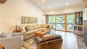 A spacious living room with a large sofa, a coffee table, and a TV. There is a balcony with a view of trees.