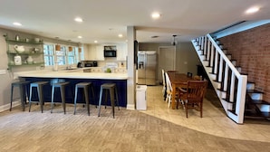 Una cocina moderna con una isla central, taburetes de bar, un comedor con mesa y sillas de madera, y una escalera que lleva a un altillo.
