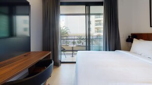 Une chambre d’hôtel équipée d’un lit, d’un bureau, d’une télévision et d’un balcon offrant une vue sur les bâtiments.
