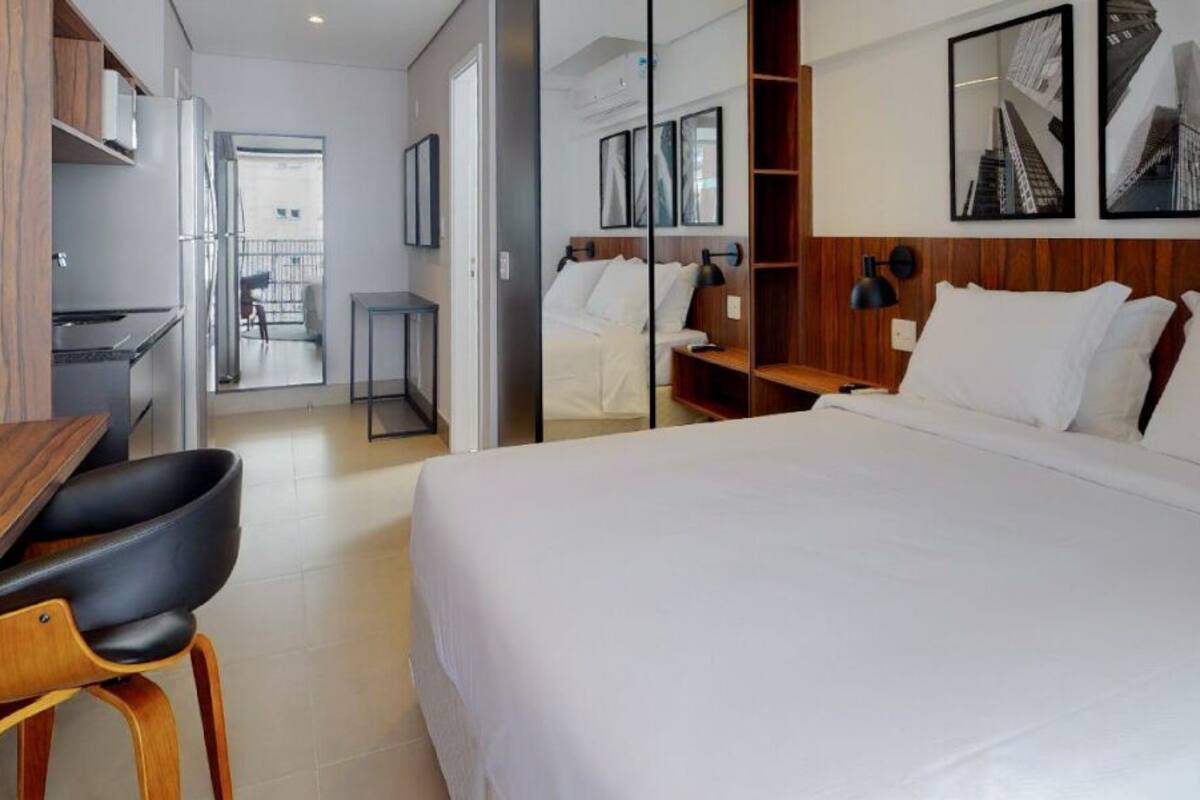 Habitación de hotel moderna con una cama grande, un escritorio con una silla, una mesita pequeña y un espejo. Se observan cuadros enmarcados en la pared y un balcón visible a través de la puerta.