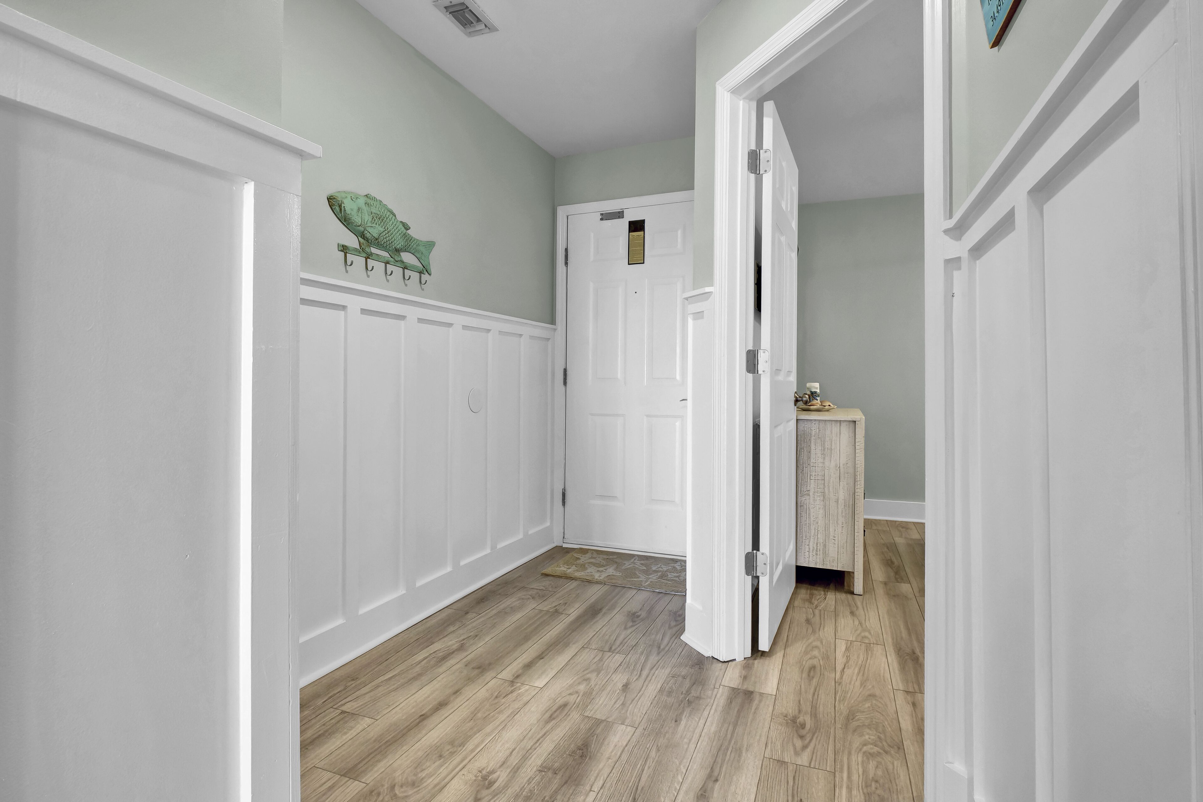 Un couloir avec des lambris blancs, un plancher en bois et une porte fermée, devant laquelle se trouve un petit paillasson.