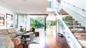 Un salon moderne avec un canapé, une table basse et des livres. Un escalier avec des rampes en verre mène à un espace extérieur.