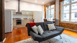 A modern living room with a grey sofa, kitchen area with white cabinets, and a large window with a view of buildings. - Toodle's Characterful loft on Lorne Street (Auckland)