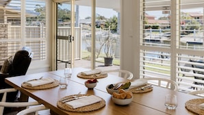 Une table à manger dressée avec des assiettes, des couverts et de la verrerie, donnant sur un quartier résidentiel grâce à de grandes fenêtres.