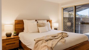 Une chambre à coucher avec une tête de lit en bois, un grand lit avec de nombreux oreillers, une table de chevet avec une lampe, et une vue sur un mur de briques à travers une porte vitrée.