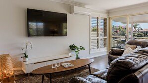 Un salon moderne avec un téléviseur à écran plat fixé au mur, un confortable canapé en cuir, une table basse en bois avec des verres et un magazine, une plante en pot et une grande fenêtre avec des stores.