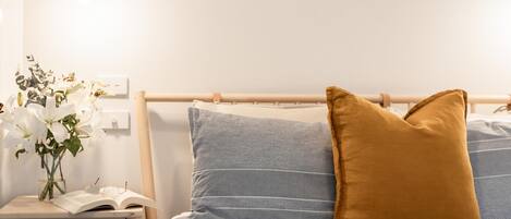 A neatly made bed with blue and mustard pillows, a bedside table with a book and glasses, and a vase with white flowers.
