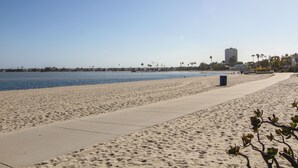A wide sandy beach with a concrete walkway, palm trees, and a distant building.