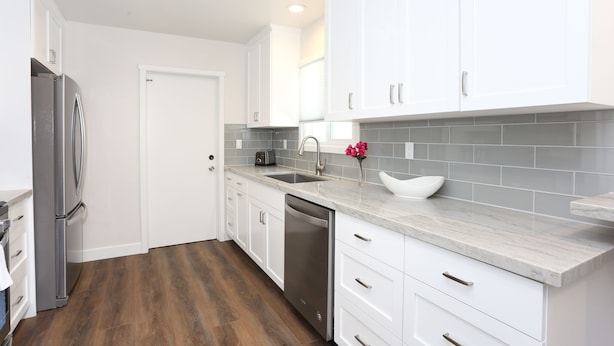 A modern kitchen with white cabinets, a stainless steel refrigerator, a dishwasher, and a marble countertop.