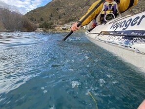 Un bote con el nombre "Voyager" en el costado, una persona remando y un paisaje montañoso al fondo.