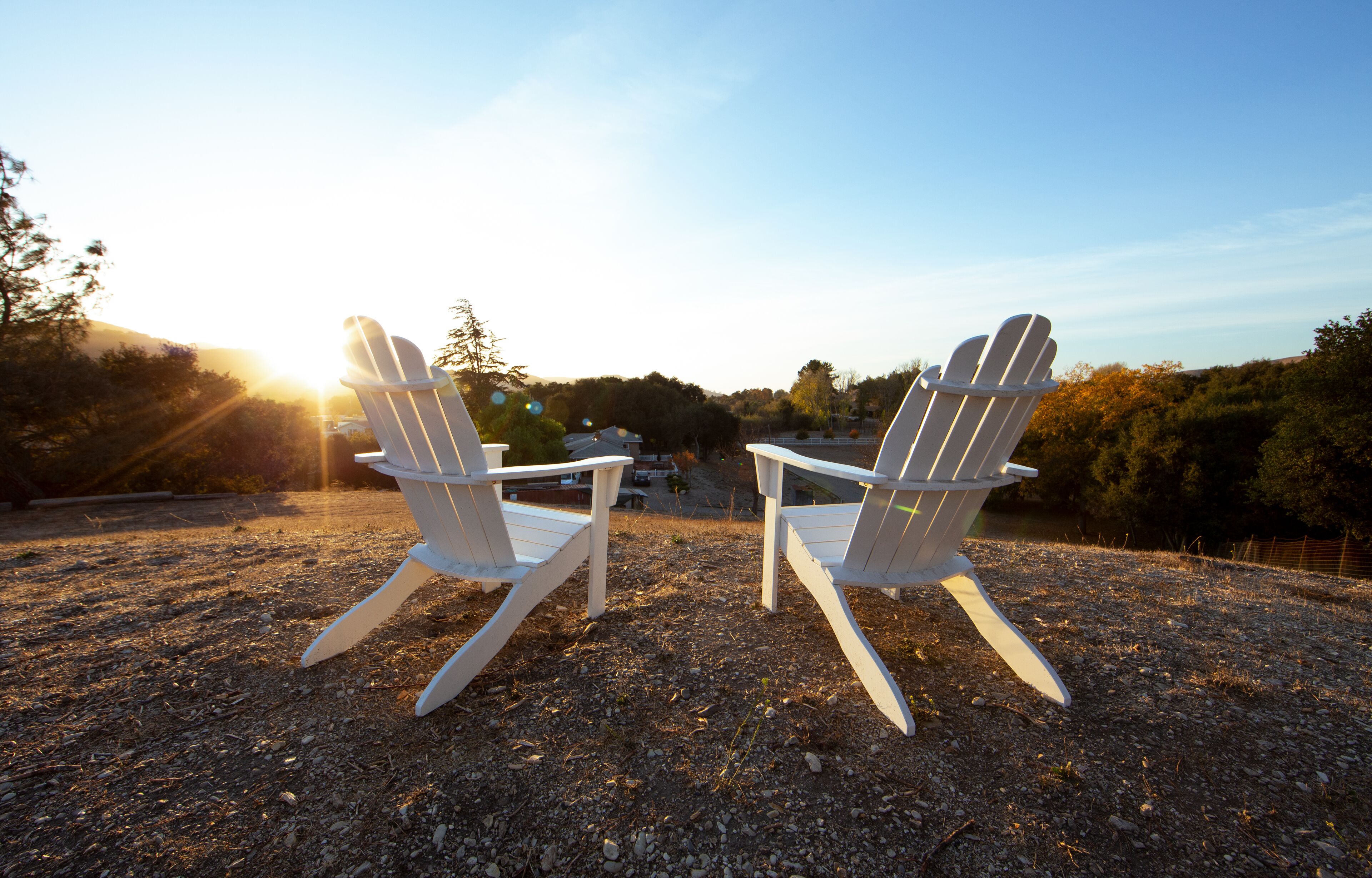 Two white Adirondack chairs facing each other on a gravel surface with a sunset in the background.