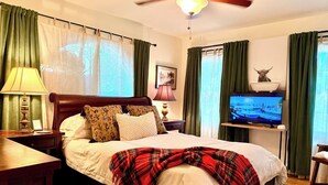 Una habitación de hotel con cama, televisión, ventilador de techo y ventana con cortinas.