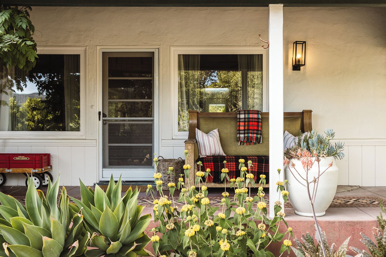 A porch with a bench, a red wagon, and a large potted plant.
