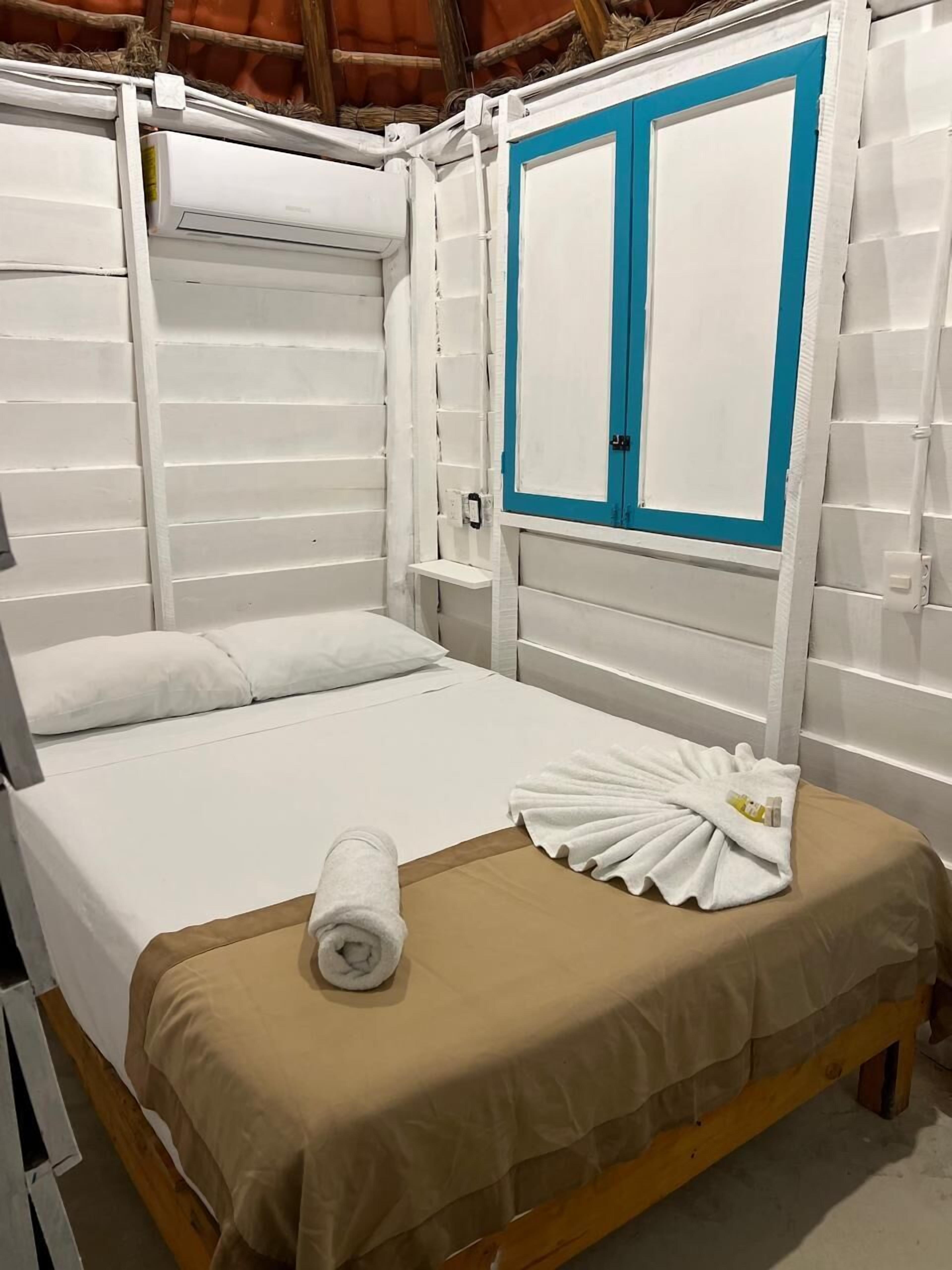 A single bed with a beige bedspread, a white towel, and a blue-framed wardrobe in a room with white walls and wooden beams.