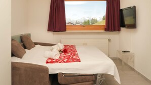 Une chambre à coucher bien rangée, avec un lit, des oreillers, une table de chevet, un téléviseur et une fenêtre avec des rideaux.