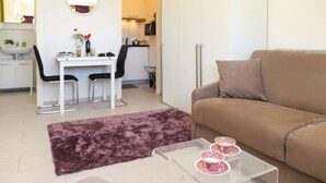 Un salon moderne avec un canapé marron, une table basse en verre agrémentée d’objets décoratifs, un coin repas avec une table et des chaises blanches, et une cuisine en arrière-plan.