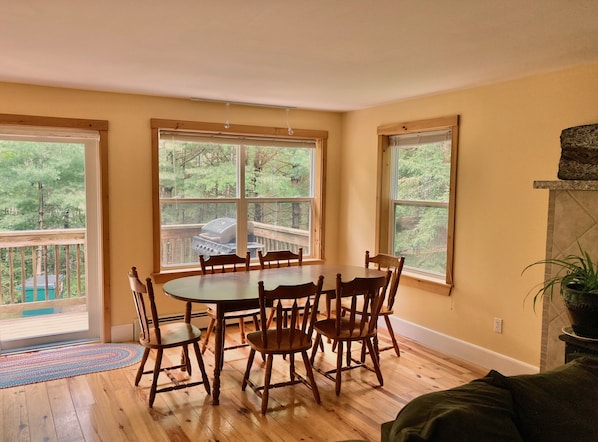 A dining area with a wooden table and chairs, large windows with a view of a deck and trees, and a fireplace.