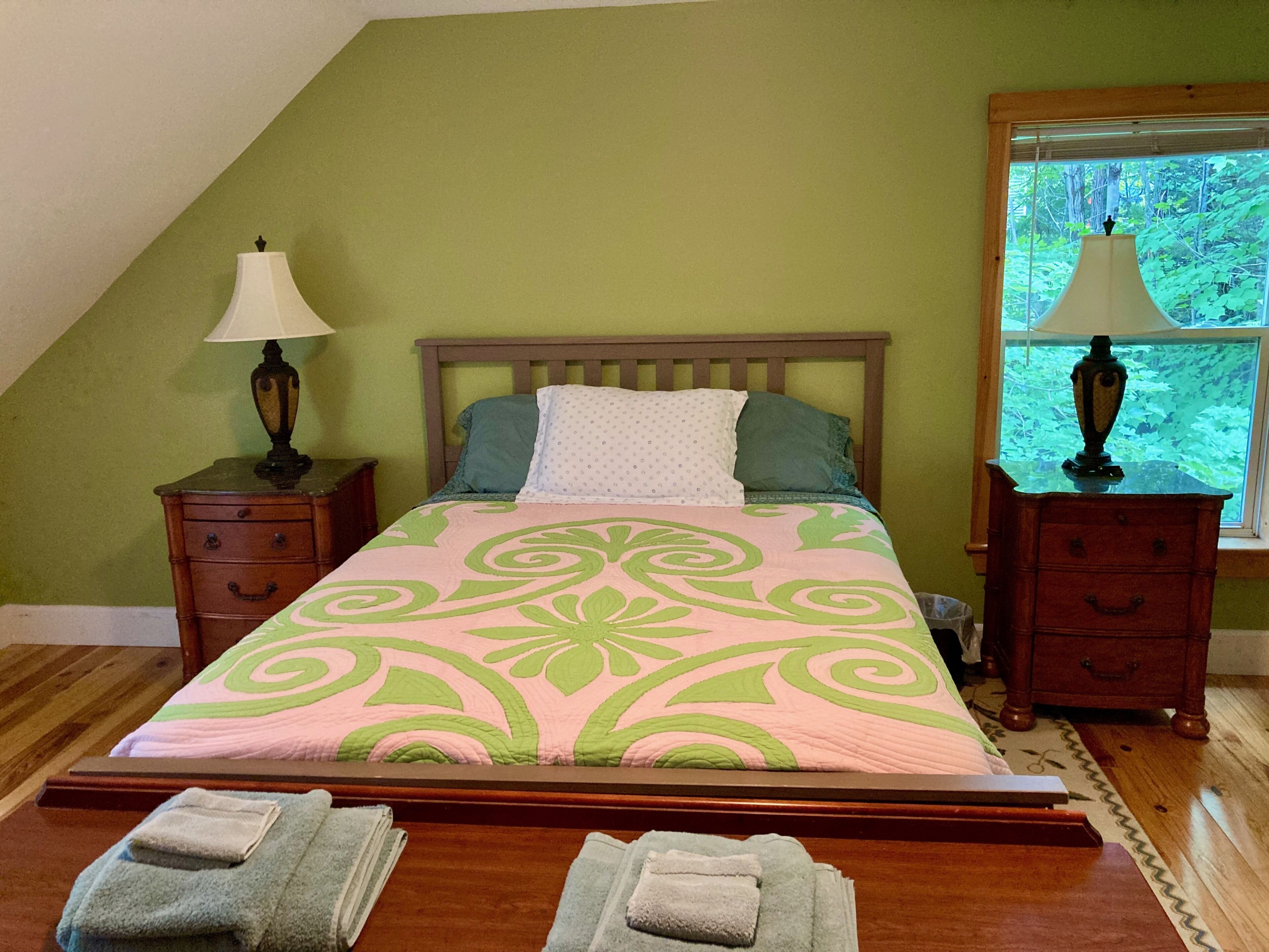A bedroom with a bed, two nightstands, a window, and a view of greenery outside.