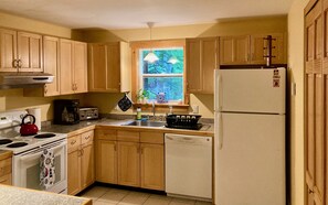A kitchen with wooden cabinets, a stove, a refrigerator, a sink, and a window with a view of trees.