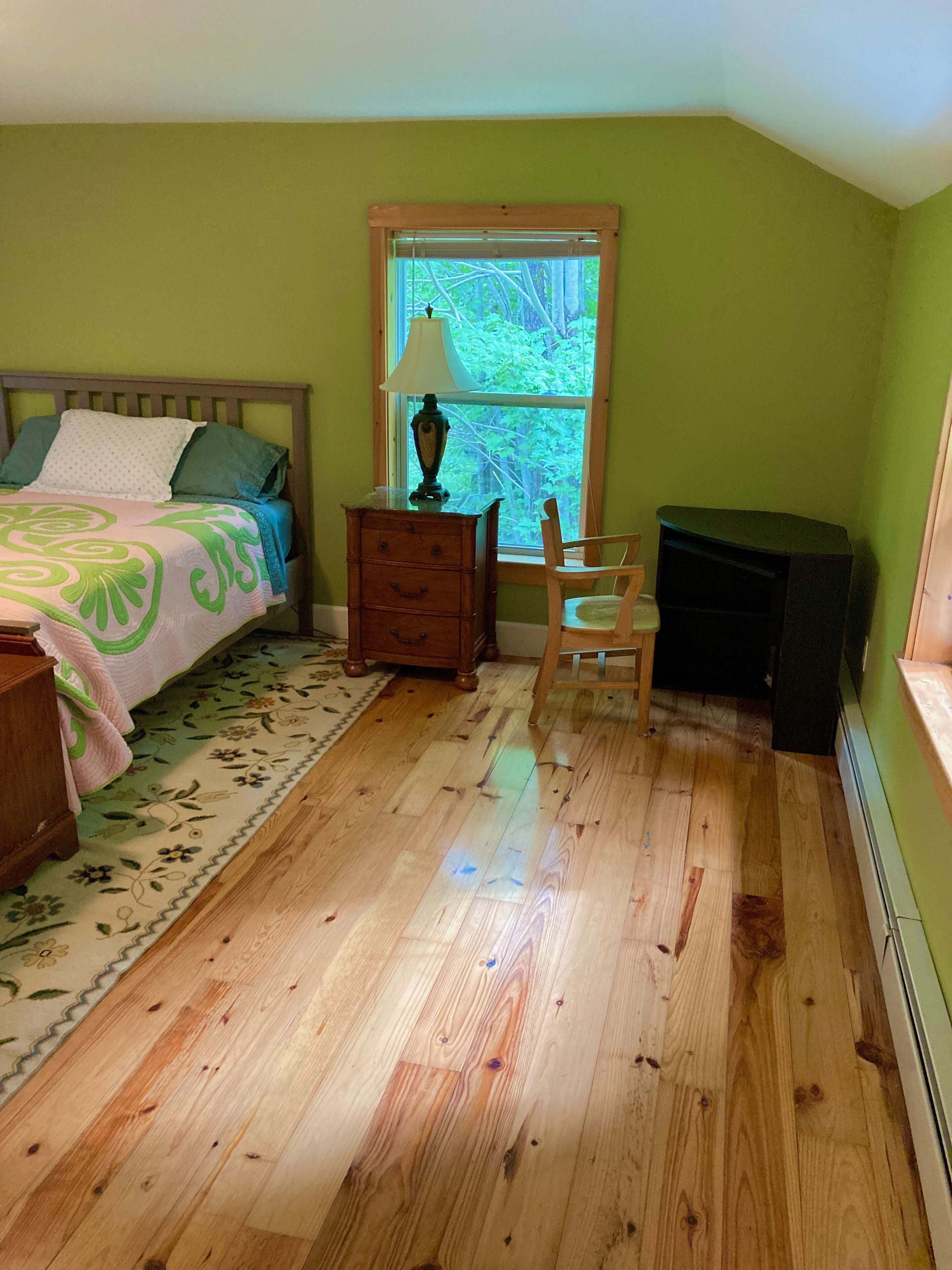 A bedroom with a bed, a nightstand, a chair, a lamp, and a window with a view of trees.