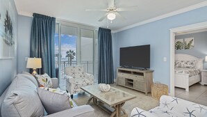 Un salon comprenant un canapé, un fauteuil, une table basse, un téléviseur et un balcon avec des rideaux.