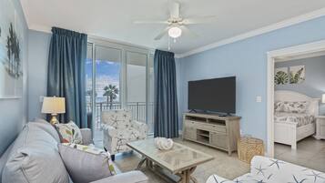 En stue med sofa, lænestol, sofabord, tv og en balkon med gardiner.