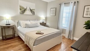 A bedroom with a bed, two bedside tables, a nightstand, a ceiling fan, a window with curtains, and a wall with framed pictures. - Luxury Ocean View Getaway! (Jacksonville Beach)
