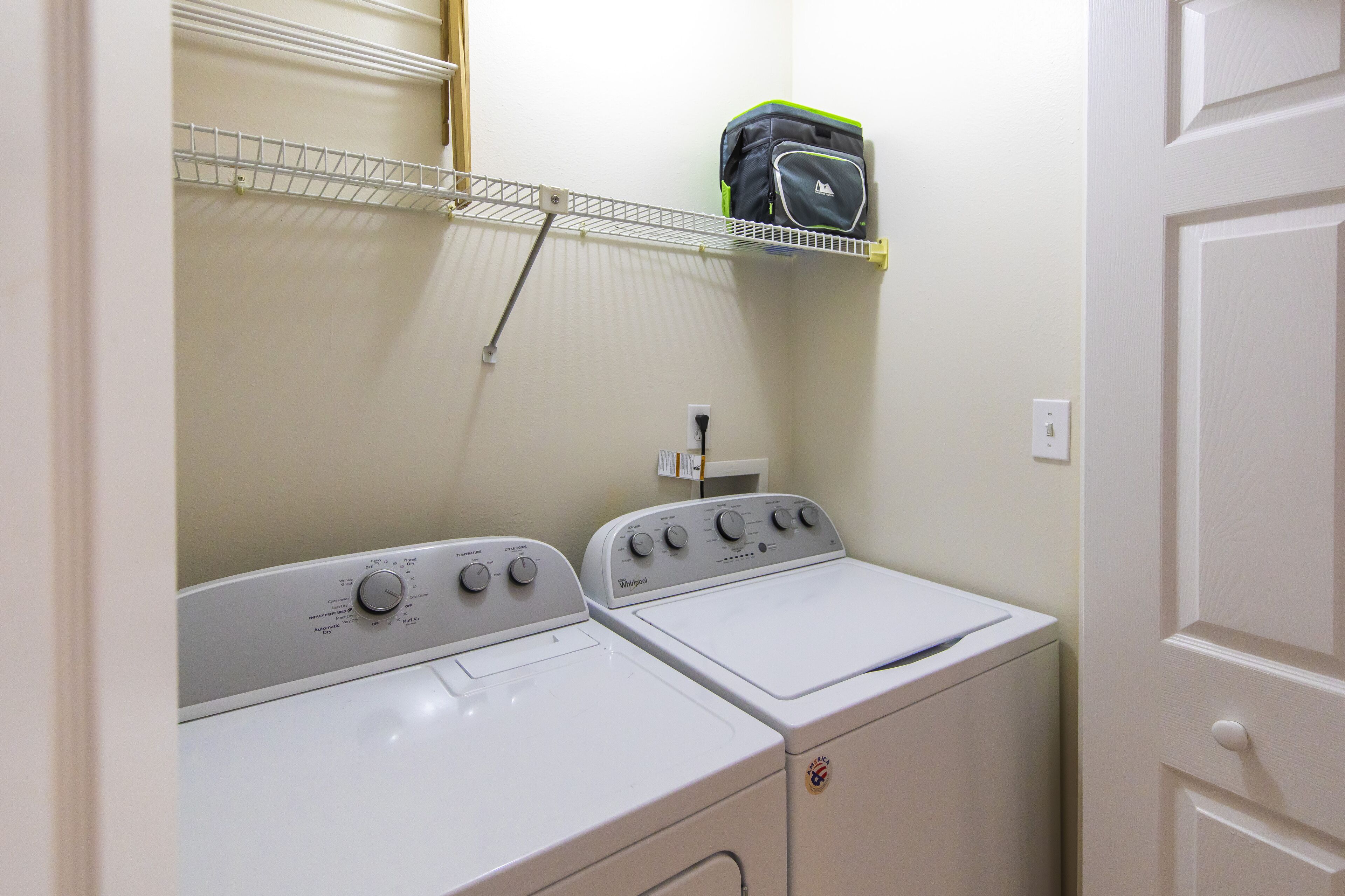 A laundry area with two white Whirlpool washers and dryers, a metal shelf above, and a door to the right.